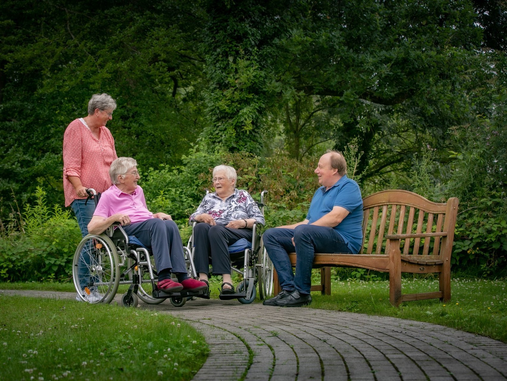 Vier Personen unterhalten sich in einem grünen Park, eine Person sitzt auf einer Holzbank.