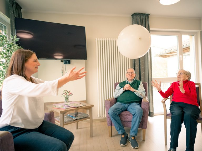 Eine junge Frau und zwei Senioren spielen in einem hellen Raum. Die Frau links und die ältere Dame rechts werfen sich einen weißen Ballon zu, während ein älterer Herr in der Mitte zuschaut.