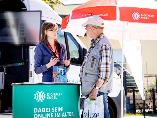 Eine Frau berät an einem grünen Informationsstand mit dem Schriftzug "Digitaler Engel" einen älteren Mann. Sie gestikuliert beim Sprechen und hält Broschüren. Im Hintergrund sind ein weißer Van und ein rot-weißer Sonnenschirm zu erkennen.