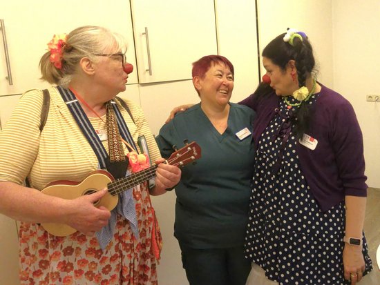 Drei fröhliche Frauen stehen zusammen. Zwei tragen rote Clownsnasen und Blumen im Haar; die linke Frau hält eine Ukulele. Die Frau in der Mitte trägt medizinische Kleidung. Sie lachen gemeinsam.