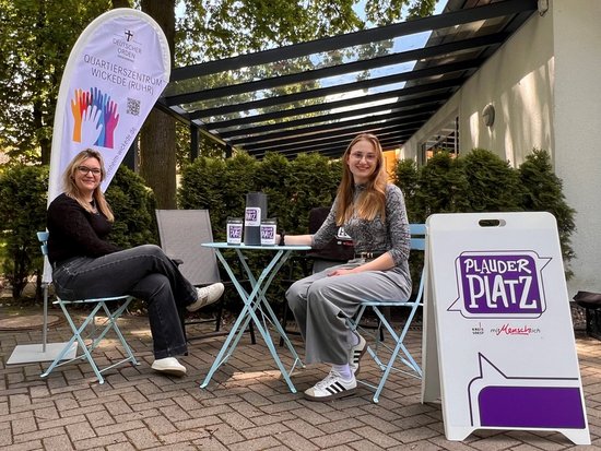 Zwei Frauen sitzen lächelnd auf Stühlen an einem kleinen Tisch im Freien. Rechts steht ein Schild mit der Aufschrift „Plauder Platz“ und links eine Fahne mit bunten Handabdrücken. Auf dem Tisch liegen Informationsmaterialien mit QR-Codes.