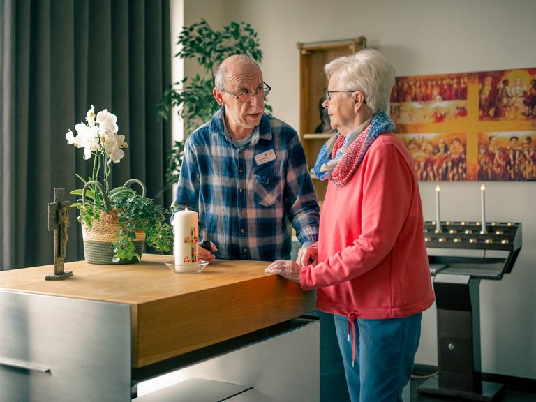 Ein älterer Mann und eine ältere Frau steht vor einem Altar in einer Kirche und unterhält sich. Der Mann trägt ein kariertes Hemd, die Frau eine rosa Jacke. Auf dem Altar stehen Kerzen und ein Kreuz.