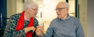 Ein älteres Paar in einem Flur. Eine Frau mit rotem Schal hält die Hand eines Mannes im Rollstuhl, während sie sich anlächeln und unterhalten.